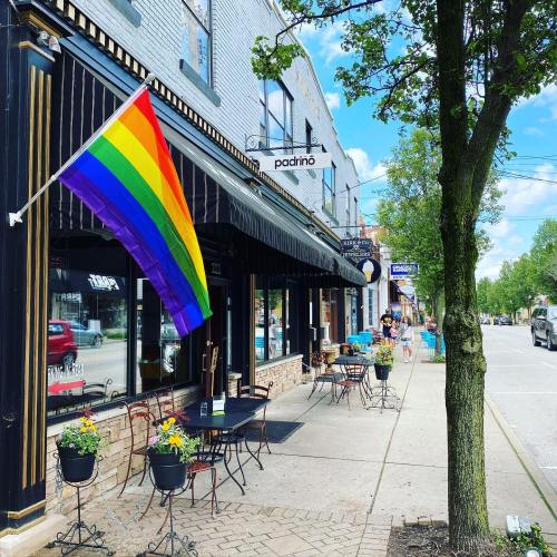 A colorful rainbow flag hangs outside a restaurant, with outdoor seating and trees lining the street on a sunny day.