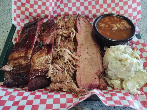 A platter featuring ribs, pulled pork, brisket, baked beans, and potato salad on a checkered paper-lined tray.
