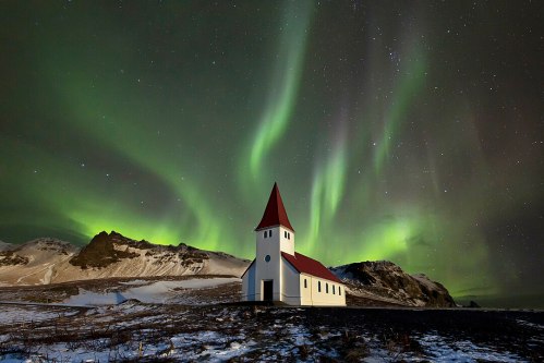 A white church with a red roof under a vibrant green aurora borealis and starry night sky. Snowy mountains in the background.