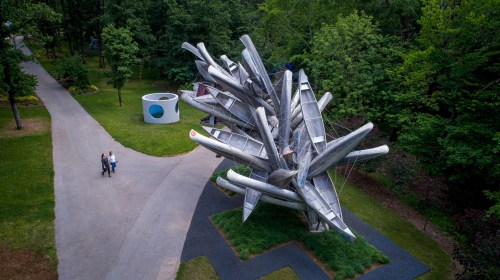 Aerial view of a large, abstract sculpture in a park, with visitors admiring it along a pathway surrounded by trees.