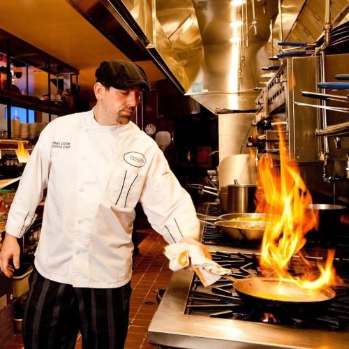A chef in a kitchen skillfully handles a flaming pan, showcasing culinary expertise amidst a professional setting.