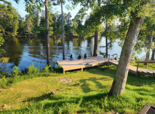 Two people sit on a wooden dock by a calm river, surrounded by lush greenery and tall trees under a clear sky.