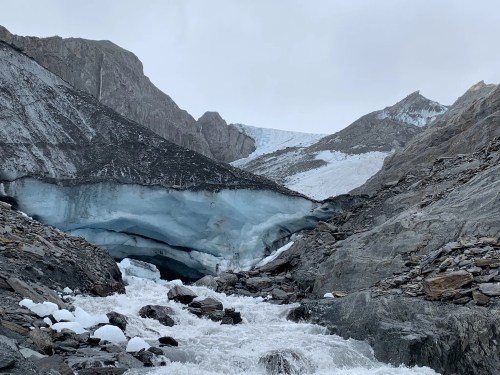 A glacial landscape with icy blue formations, rocky terrain, and a flowing river under a cloudy sky.