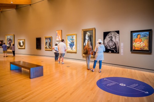 Visitors explore an art gallery with various paintings displayed on the walls and a wooden bench in the center.