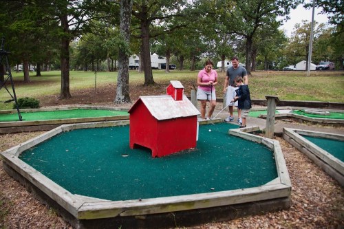 A family plays mini-golf on a green course with a red house structure in the center, surrounded by trees.