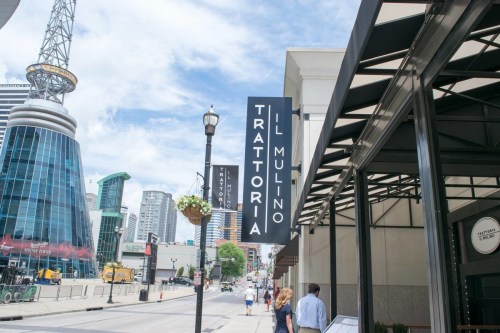 A street view featuring a restaurant sign for "Trattoria Il Mulino" with modern buildings in the background.