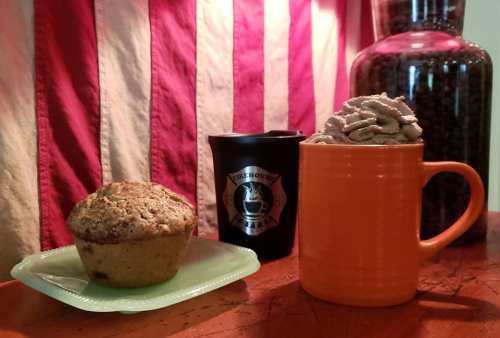 A muffin on a green plate next to a black cup and an orange mug filled with whipped cream, set against a striped background.