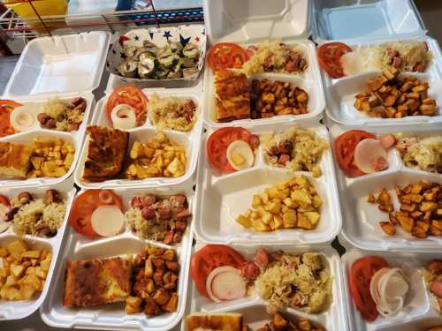 A variety of takeout food containers filled with sausages, potatoes, tomatoes, and sauerkraut arranged on a table.