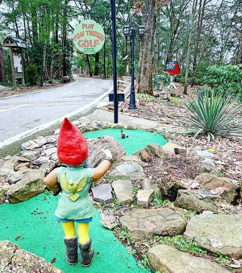 A whimsical gnome statue stands on a green path, with a sign for mini golf in a wooded area.