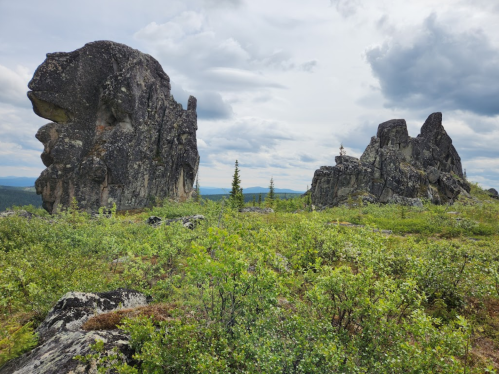 Two large rock formations rise from a green landscape under a cloudy sky, surrounded by sparse vegetation.