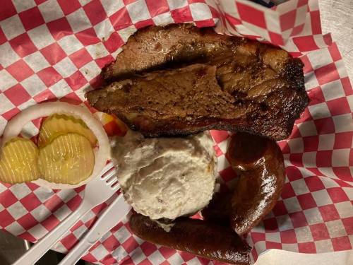 A plate of barbecue featuring brisket, sausage, mashed potatoes, pickles, and onions on a red and white checkered paper.