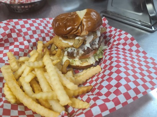 A crispy chicken sandwich with pickles and sauce on a pretzel bun, served with a side of crinkle-cut fries in a red checkered basket.
