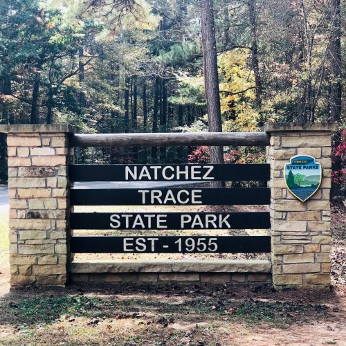 Sign for Natchez Trace State Park, established in 1955, surrounded by trees in a wooded area.