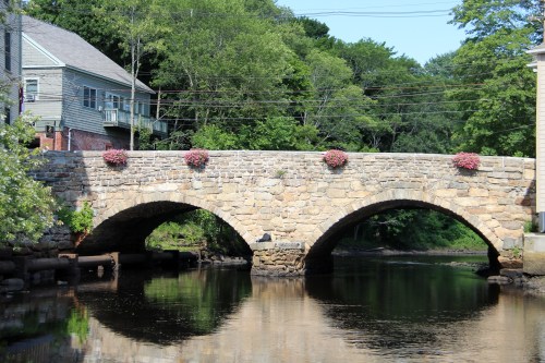A stone bridge with flower pots spans a calm river, surrounded by greenery and buildings on either side.