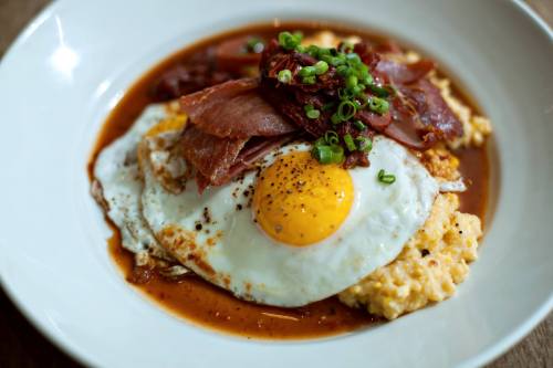 A bowl of creamy grits topped with a fried egg, crispy bacon, and green onions, drizzled with a savory sauce.