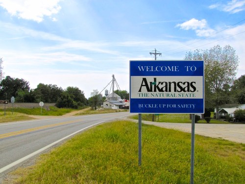 Welcome sign for Arkansas, featuring "The Natural State" and a reminder to buckle up for safety, with a rural backdrop.