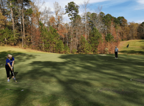 Two golfers on a green, one preparing to putt while the other watches, surrounded by autumn trees.