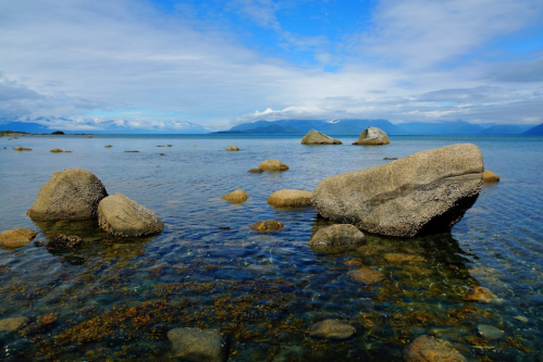 A serene lake scene with smooth rocks emerging from calm waters under a cloudy blue sky.