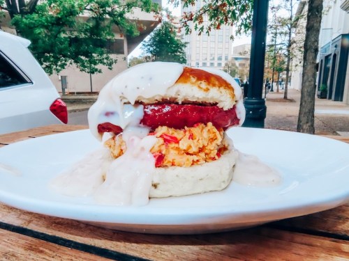 A close-up of a hearty sandwich with sausage, chicken, and creamy sauce on a biscuit, served on a white plate outdoors.