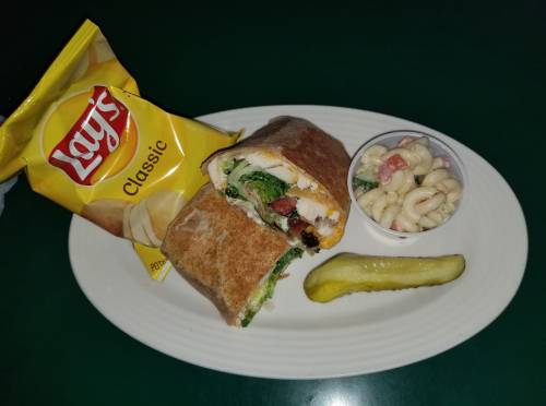 A plate with a wrap filled with greens, a side of macaroni salad, a pickle, and a bag of Lay's Classic chips.