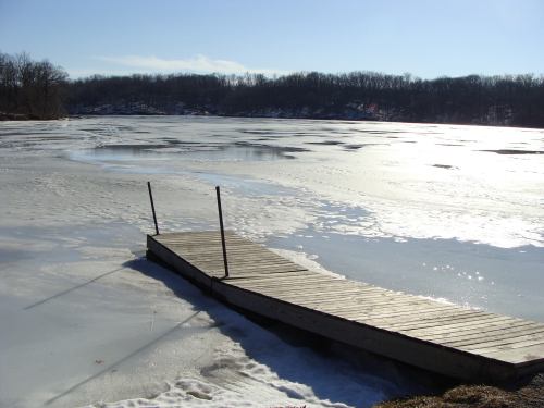 A wooden dock extends over a frozen lake, surrounded by trees and a clear blue sky.