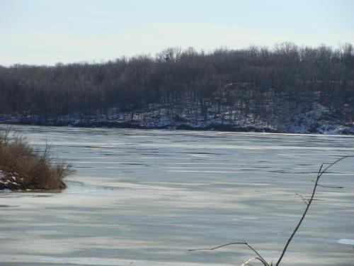 A frozen lake surrounded by a snowy landscape and bare trees under a clear blue sky.