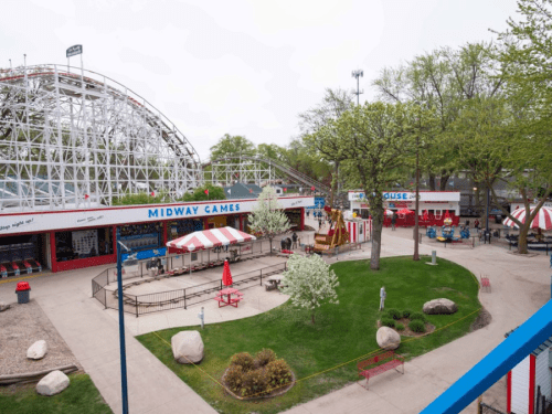 A view of an amusement park featuring a roller coaster, game stalls, and trees in a green, open area.