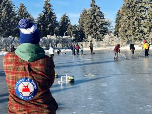 A person in a plaid jacket watches a curling game on an outdoor rink, surrounded by trees and other players.