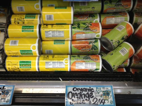 Cans of organic orange juice and lemonade stacked on a store shelf, with a price tag for orange juice.