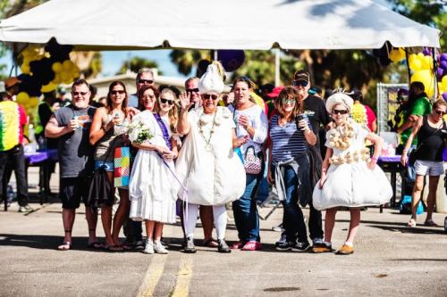 A lively group of people in festive attire poses under a tent at an outdoor event, holding drinks and smiling.