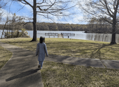 A child walks along a path by a lake, with trees and a dock visible in the background under a clear sky.
