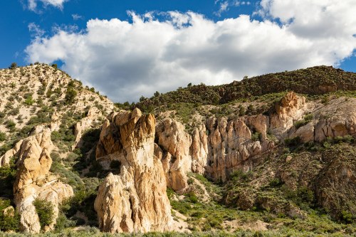 Rocky cliffs and formations under a blue sky with fluffy clouds, surrounded by green vegetation.