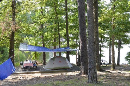 A campsite with a tent, picnic table, and blue tarp, surrounded by trees near a lake. A bicycle is nearby.