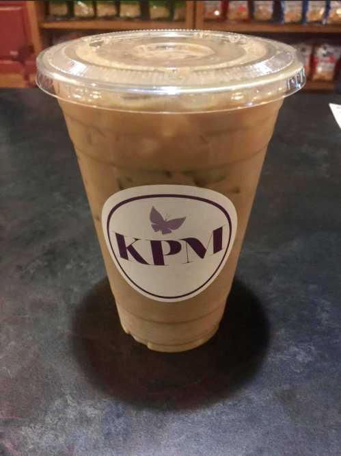 A clear cup of iced coffee with a logo labeled "KPM" on a dark countertop.