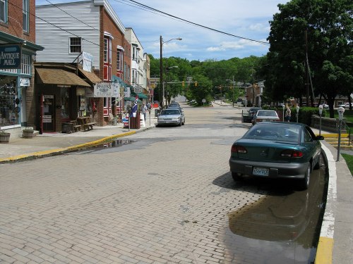 A street scene with shops, parked cars, and a puddle on the cobblestone road under a blue sky with scattered clouds.