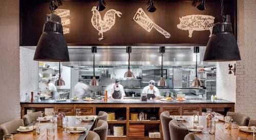 A modern restaurant interior with a visible kitchen, wooden tables, and chefs preparing food behind a glass partition.