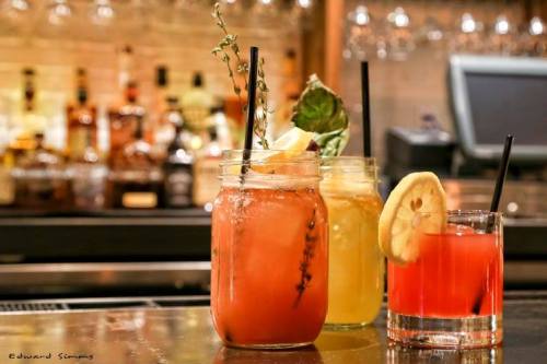 Three colorful cocktails in jars, garnished with herbs and citrus, set against a blurred bar background.