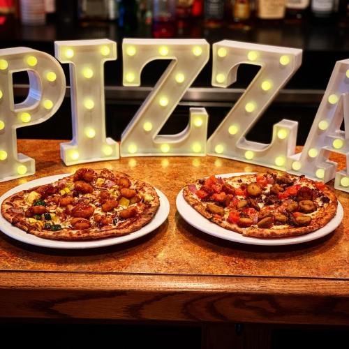 Two pizzas topped with various ingredients sit on a wooden table, with illuminated letters spelling "PIZZA" in the background.