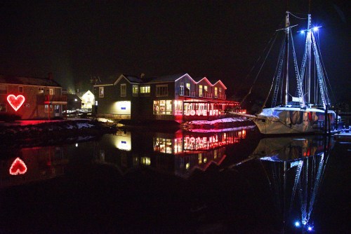 A nighttime scene of a harbor with a lit-up building and a boat, reflecting in calm water, featuring a heart decoration.
