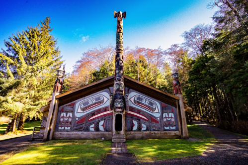 A traditional totem pole stands in front of a wooden structure, surrounded by tall trees and a clear blue sky.