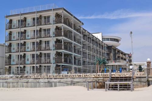 A modern beachfront hotel with multiple balconies and a pool area, surrounded by sandy beach and blue sky.