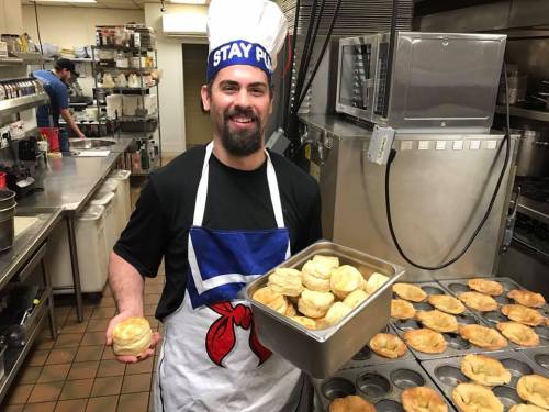 A smiling chef in an apron holds a tray of freshly baked biscuits in a busy kitchen.