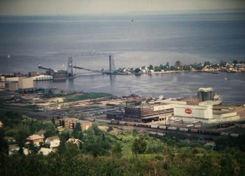Aerial view of a harbor with a bridge, industrial buildings, and water, surrounded by greenery and distant shoreline.