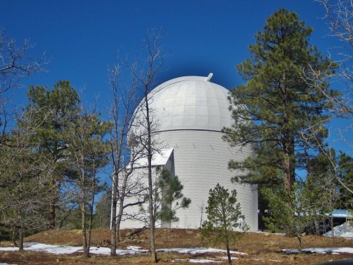 A white observatory dome surrounded by pine trees and snow under a clear blue sky.