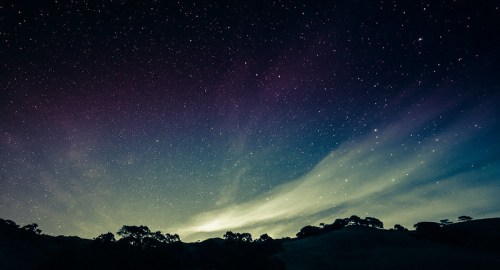 A starry night sky with colorful hues and wispy clouds above a dark silhouette of trees and hills.