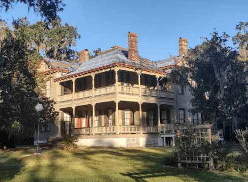 A large, historic house with multiple balconies, surrounded by trees and greenery under a clear blue sky.