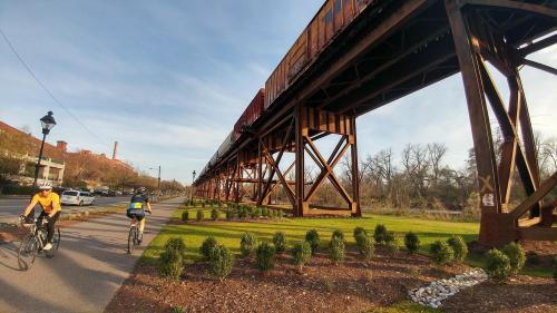 Two cyclists ride along a path beneath a large elevated train track, with greenery and buildings in the background.
