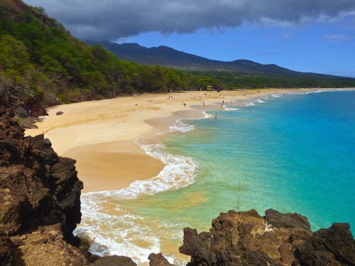 A scenic beach with golden sand, turquoise water, and lush green hills under a partly cloudy sky.
