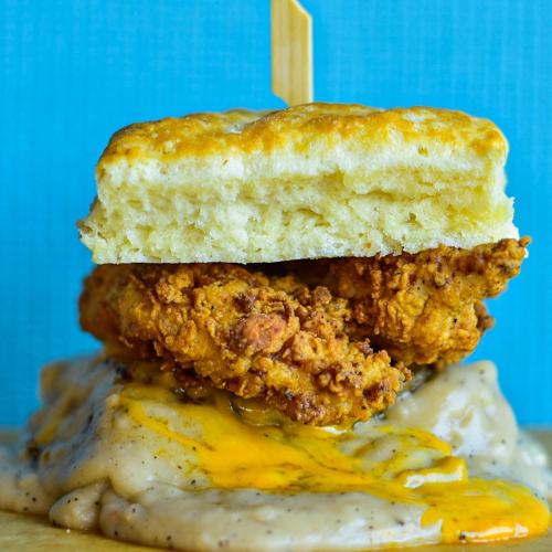 A close-up of a biscuit sandwich with fried chicken, drizzled with creamy gravy and topped with a wooden skewer.