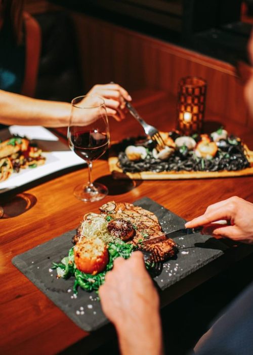 A person cuts into a steak on a slate plate, with wine and another dish in the background, in a cozy dining setting.